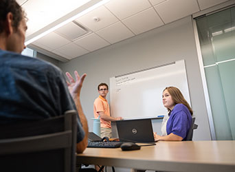 A male student stands in front of a whiteboard and a female student sits in front of a laptop while both listen to another student speak.