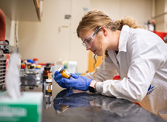 A female student in lab safety gear closely inspects a labeled bottle at her lab desk.