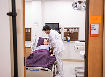 A male student in white medical scrubs uses a stethoscope on a human replica as he practices an examination in a simulation hospital room.