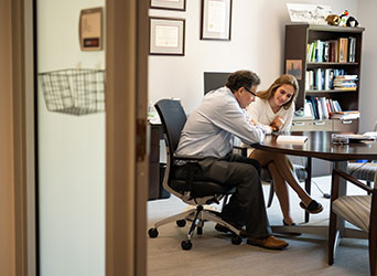 A male professor and female student sit together at a table in an office as they both read over passages in the same book.