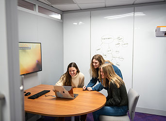 Three female students laugh as they stare at the same computer screen while they work together in a meeting room.